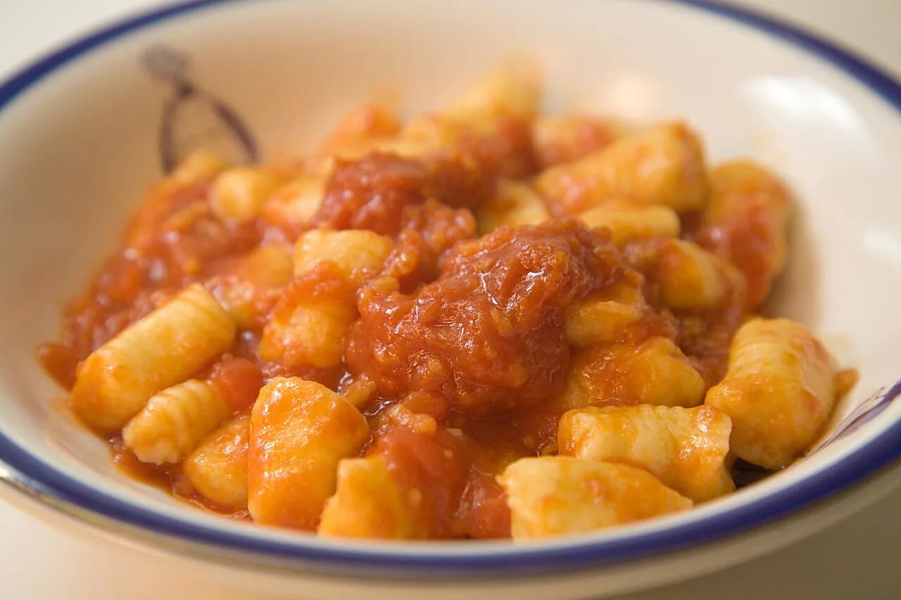 Foto da receita: Nhoque de Batata com Molho de Tomate - Nhoque macio de batata cozida, farinha e ovo, modelado à mão e cozido em água fervente até boiar. Servido com molho de tomate caseiro e parmesão. Tradição do dia 29.