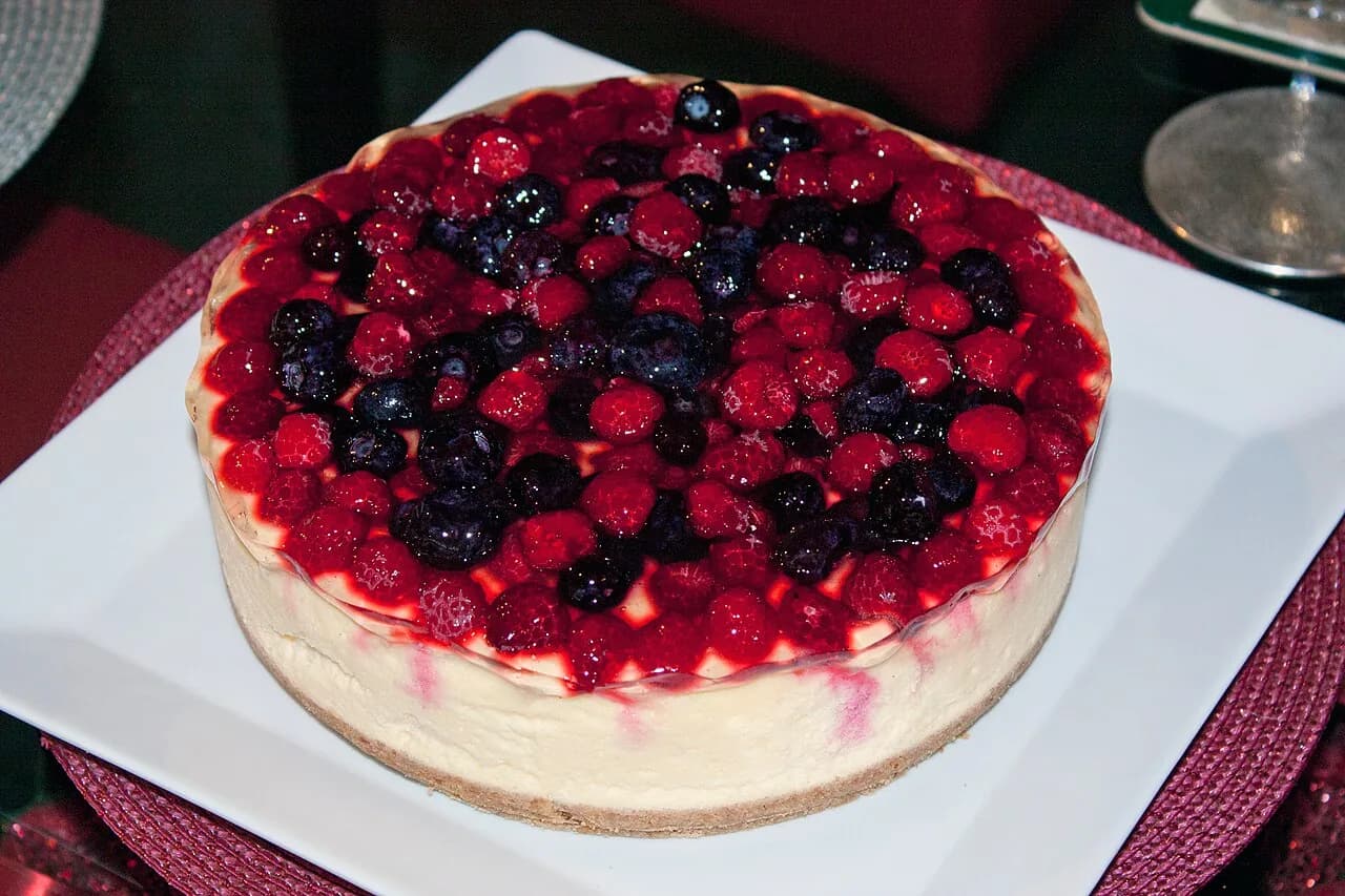 Foto da receita: Cheesecake de Frutas Vermelhas - Cheesecake assado de cream cheese com base de biscoito e cobertura de calda de morango e framboesa. Textura cremosa, sem rachaduras, no estilo nova-iorquino.
