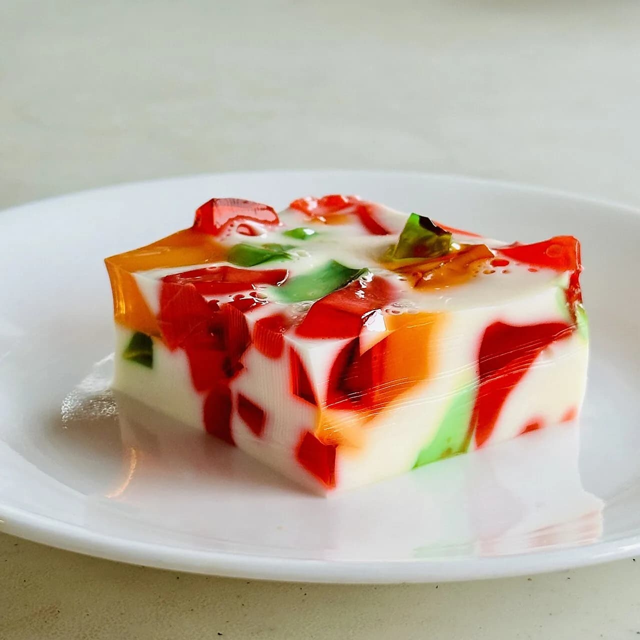 Foto da receita: Gelatina Colorida com Creme (Mosaico) - Cubos de gelatina de sabores variados envoltos em um creme branco de leite condensado que firma na geladeira. Sobremesa colorida de festa infantil.